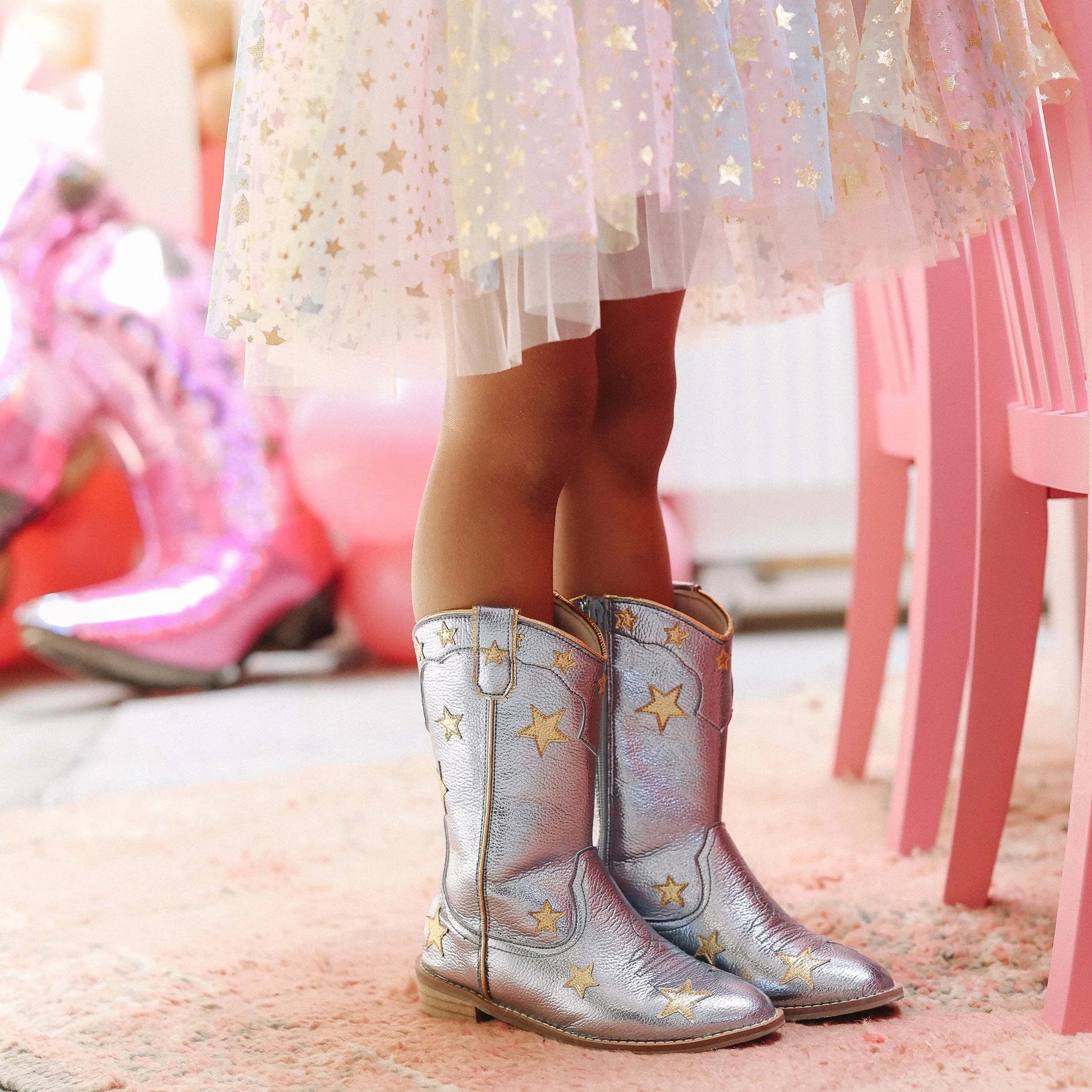 Child wearing silver cowboy boots with star patterns in a bright, possibly indoor setting.