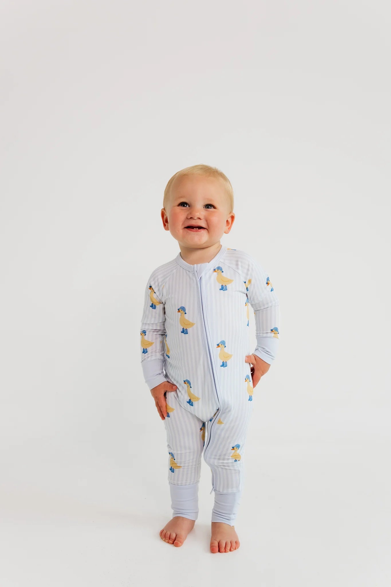 A baby wearing a light blue and white striped zip-up footless sleeper with a yellow cartoon duck print, standing against a plain white background.