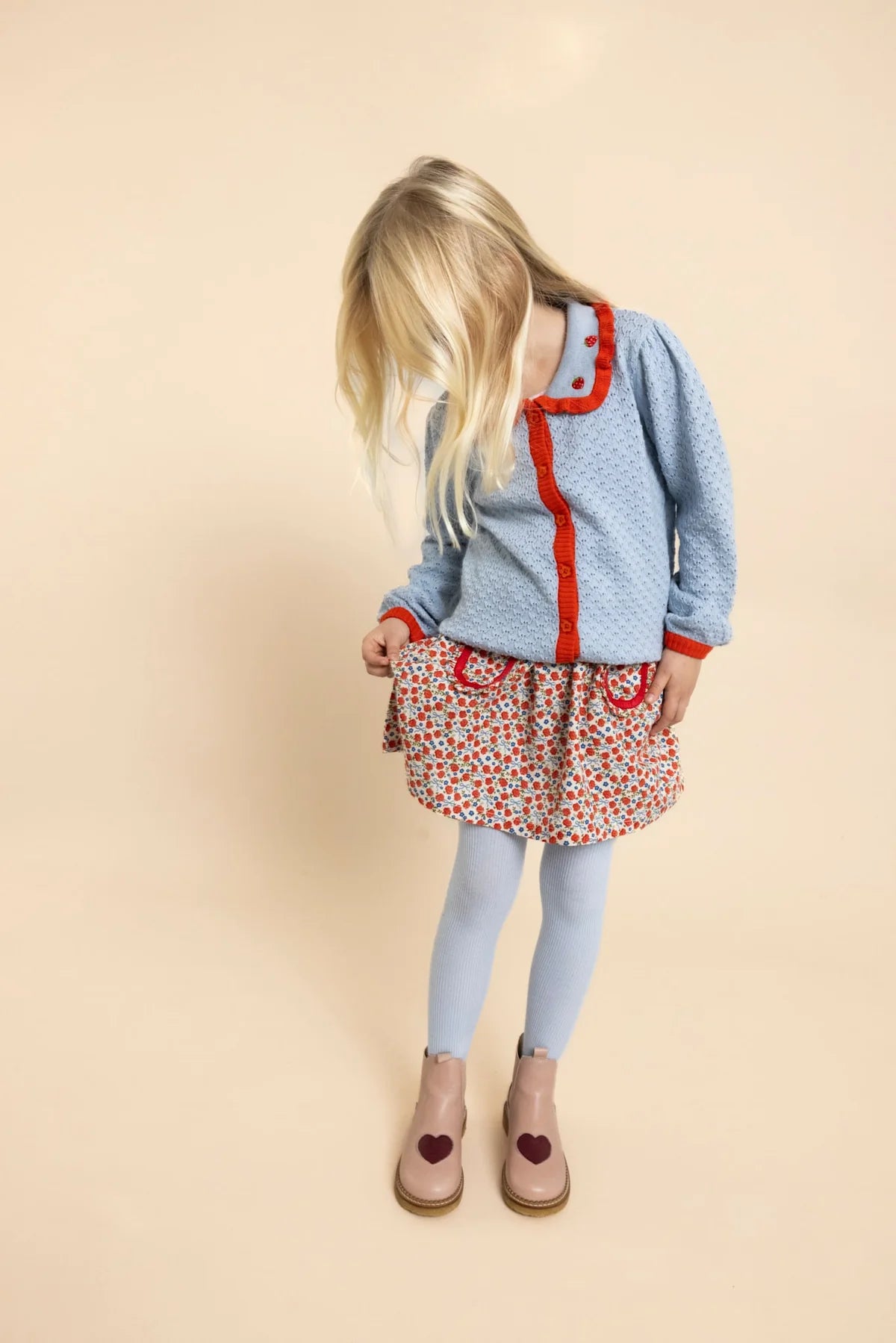 A young girl with blonde hair is shown from the back, wearing a light blue patterned cardigan with red trim, a red and white floral dress, light blue tights, and beige shoes with heart details, set against a solid beige background.