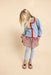 A young girl with blonde hair is shown from the back, wearing a light blue patterned cardigan with red trim, a red and white floral dress, light blue tights, and beige shoes with heart details, set against a solid beige background.