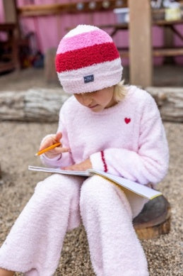 A child wearing a fuzzy pink and red striped beanie and a matching light pink fleece outfit sits outdoors on a wooden bench, writing in a notebook with a pencil.