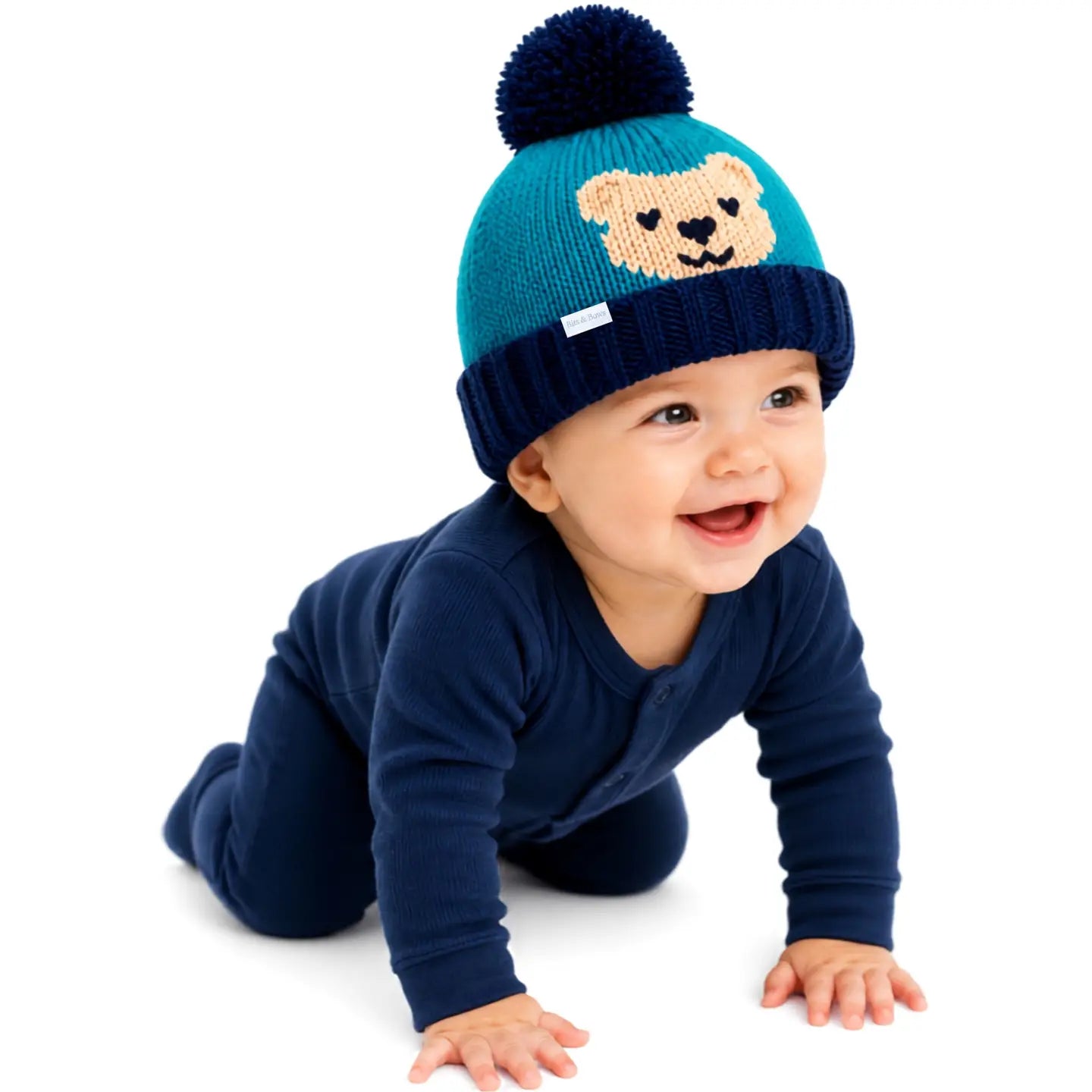 A baby crawling on a white background wearing a navy blue long-sleeve one-piece and a teal and navy knit pom-pom beanie with a teddy bear face.