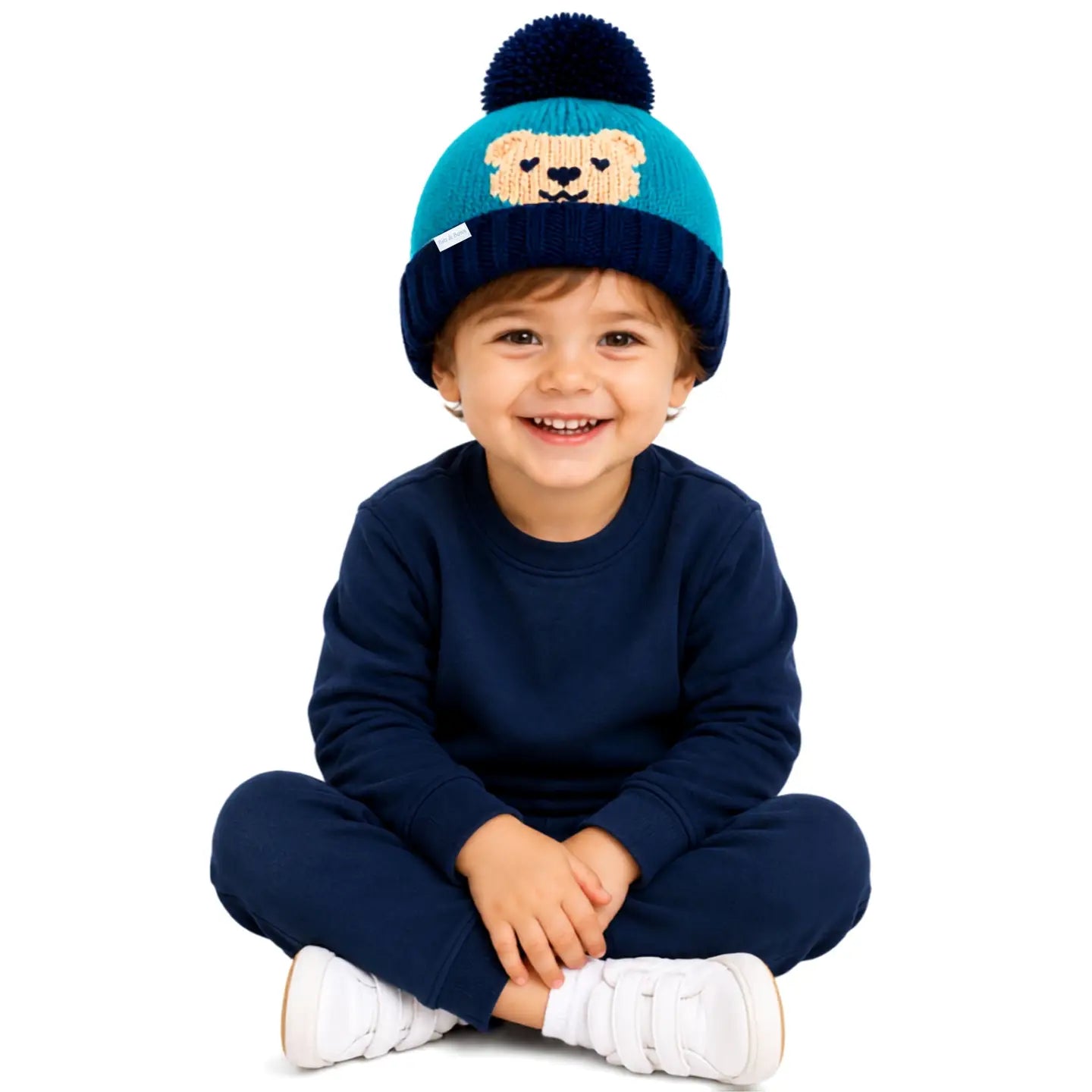 A smiling toddler sitting cross-legged on a white background, wearing a navy blue sweatshirt and pants set with a teal and navy knit pom-pom beanie featuring a dog face design.