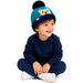 A young child sitting cross-legged on a white background, wearing a navy blue sweatshirt and matching pants, along with a teal and navy knit beanie featuring a teddy bear face and pom-pom.