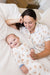 A smiling woman and baby lying on a bed, both wearing matching white pajamas with a teddy bear print and blue piping details.
