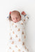 A sleeping newborn baby swaddled in a white sleep sack with a repeating pattern of small tan teddy bears wearing blue bowties, lying on a plain white background.