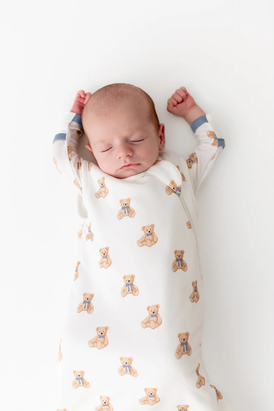 A sleeping newborn baby swaddled in a white sleep sack with a repeating pattern of small tan teddy bears wearing blue bowties, lying on a plain white background.