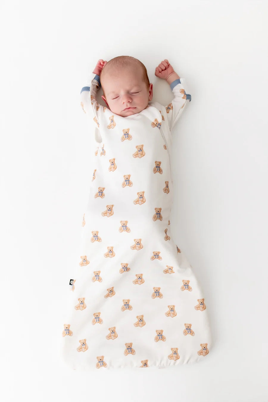 A sleeping newborn baby lying on their back, swaddled in a sleep sack printed with a repeating pattern of small tan teddy bears with blue accents, on a clean white background.