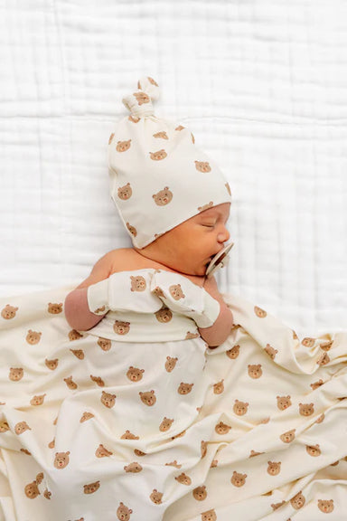 Newborn baby wrapped in a white blanket with brown bear patterns, wearing a matching white hat with brown bear patterns, on a white textured background.