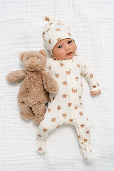 Baby in a white onesie with brown bear patterns lying on a white blanket next to a brown teddy bear.