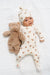 Baby in a white onesie with brown bear patterns lying on a white blanket next to a brown teddy bear.