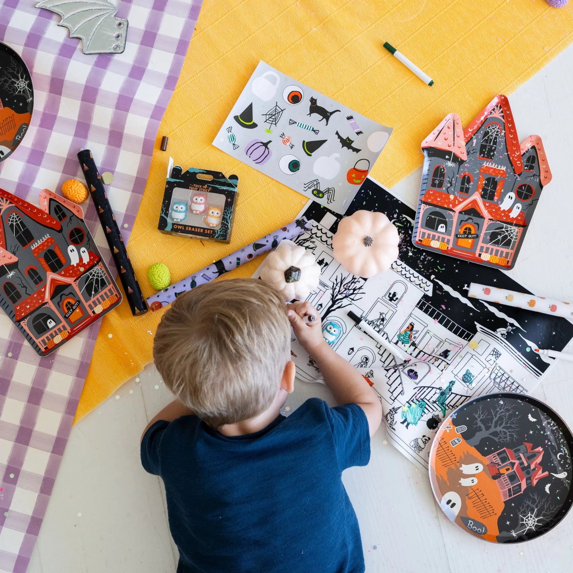 A child sits on a white floor surrounded by Halloween-themed craft supplies, including a coloring page, stickers, a small Halloween box, and decorative house cutouts, with a purple-and-white checkered blanket and yellow fabric in the background.
