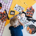 A child sits on a white floor surrounded by Halloween-themed craft supplies, including a coloring page, stickers, a small Halloween box, and decorative house cutouts, with a purple-and-white checkered blanket and yellow fabric in the background.
