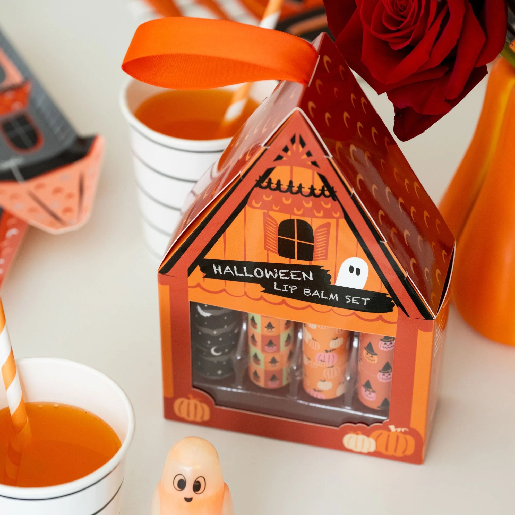 Halloween lip balm set in a house-shaped box with an orange ribbon, shown on a tabletop with cups, a rose, and small ghost decorations.