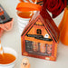 Halloween lip balm set in a house-shaped box with an orange ribbon, shown on a tabletop with cups, a rose, and small ghost decorations.