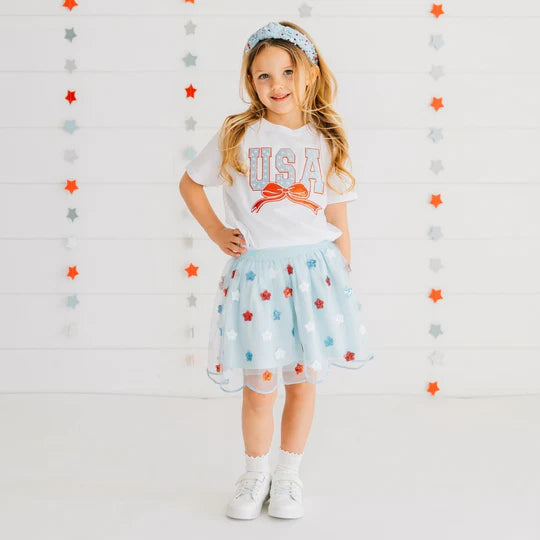 Young girl wearing a 'USA' t-shirt and star-patterned skirt against a star-patterned background