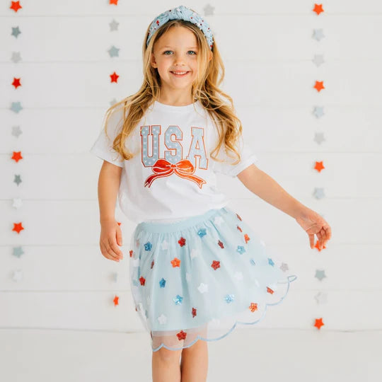 Young girl wearing a 'USA' t-shirt and light blue skirt with star patterns against a decorative background.