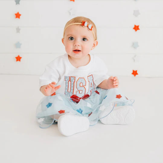 Baby sitting on a white blanket with a USA-themed outfit and headband against a star-patterned wall.