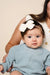 Baby wearing a light blue sweater and vanilla eyelet headband with a bow, held by an adult against a beige background.