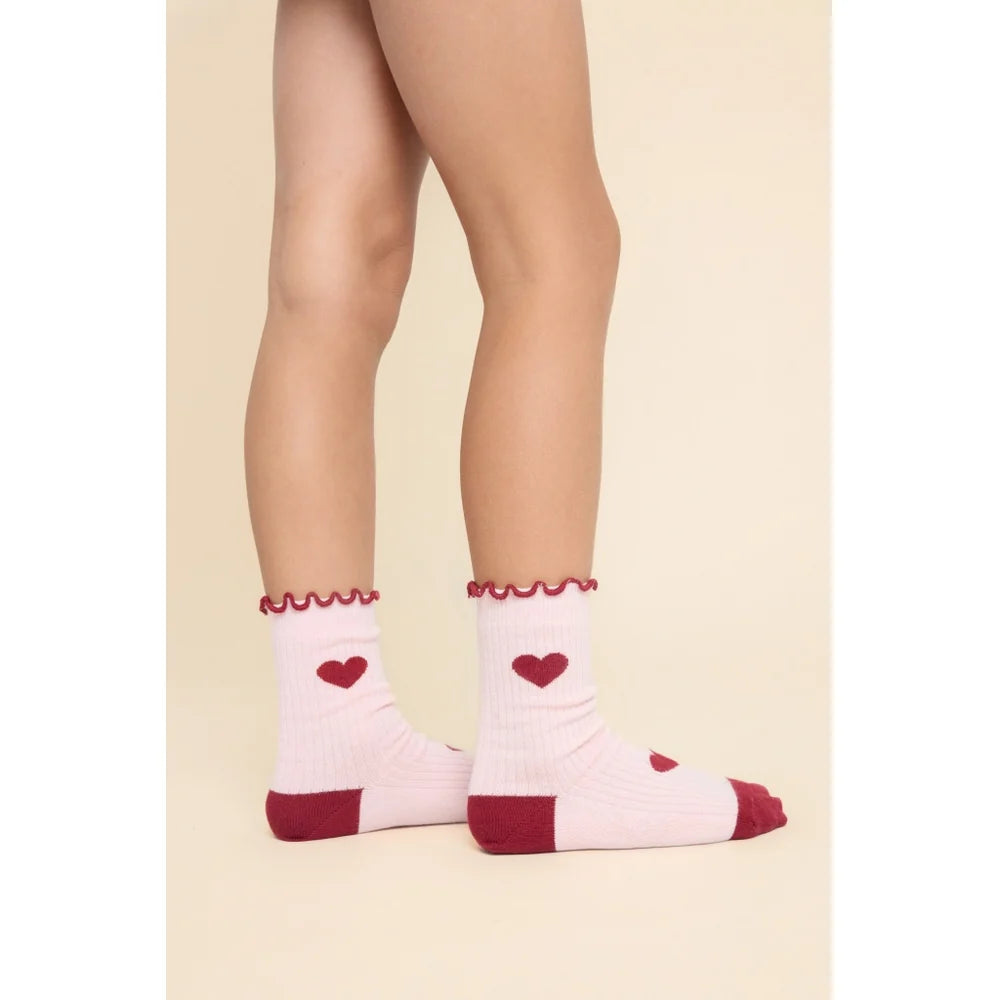 A pair of light pink crew socks with dark red heels, toes, and ruffled cuffs, featuring small red heart details on the sides, shown on a model's feet against a beige background.