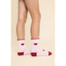 A pair of light pink crew socks with dark red heels, toes, and ruffled cuffs, featuring small red heart details on the sides, shown on a model's feet against a beige background.