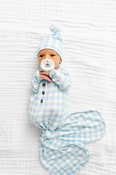 Newborn baby wrapped in a blue checkered knotted gown with a matching hat on a white textured surface.
