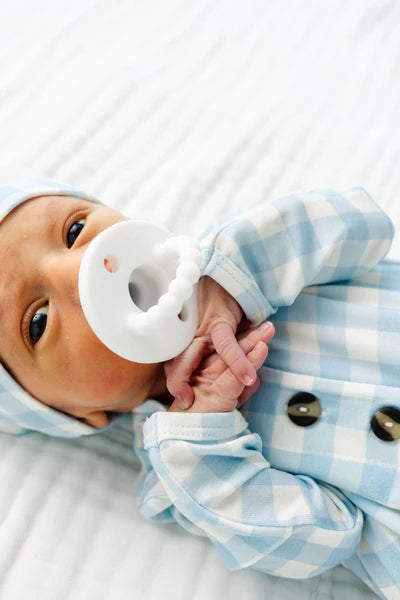 Baby wearing a blue checkered outfit with a white teething ring in mouth.
