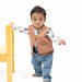 A toddler standing barefoot on a white studio background, wearing a tan fringed vest over a white long-sleeve shirt with a blue animal print, blue jeans, and holding onto a yellow chair.