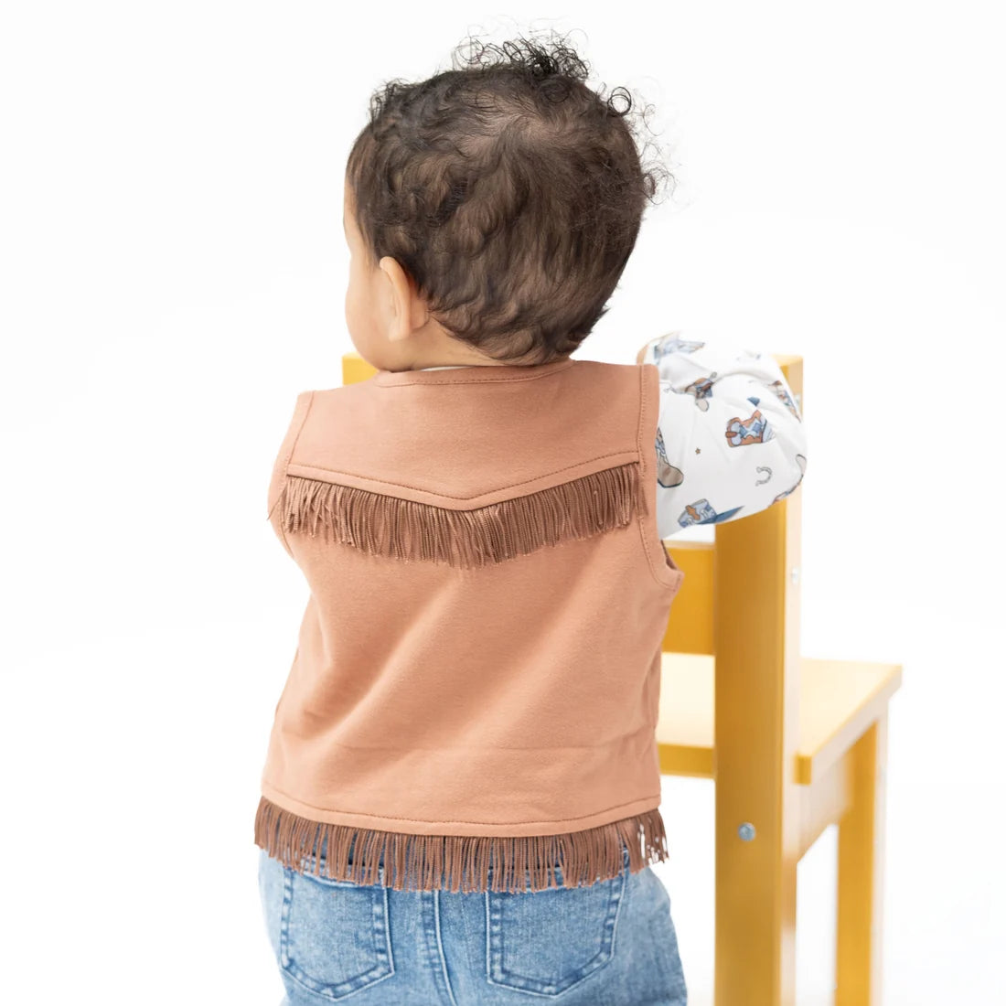 A toddler viewed from behind wearing a mocha-colored sleeveless vest with fringe trim and blue jeans, standing next to a yellow chair against a white background.