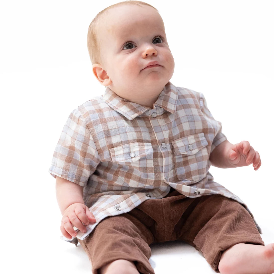 A baby sitting on a white background wearing a brown and beige plaid short-sleeve button-up shirt and brown pants.