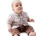 A baby sitting on a white background wearing a brown and beige plaid short-sleeve button-up shirt and brown pants.
