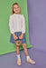 Young girl in a white eyelet shirt and patterned skirt standing against a green and purple gradient background.