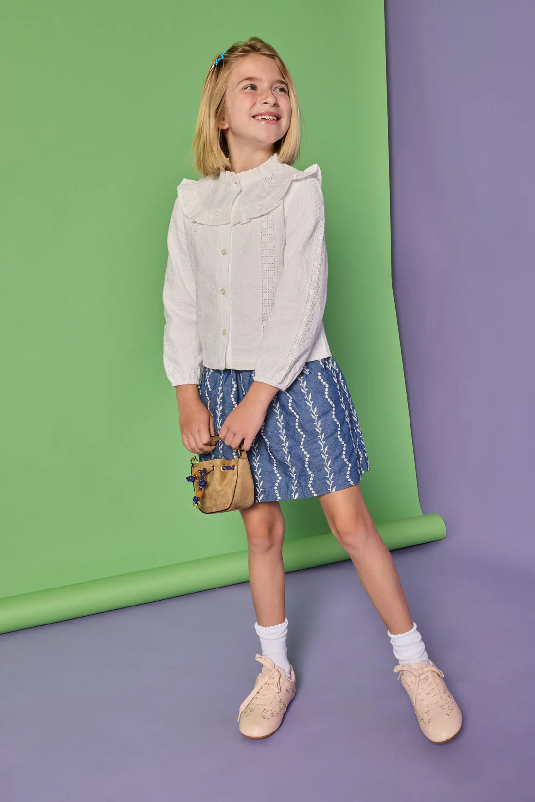A young girl stands in a studio wearing a white ruffled long-sleeve blouse, a blue patterned skirt, white socks, and beige shoes, holding a small tan handbag. The background is a two-tone green and purple seamless backdrop.