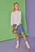 A young girl stands in a studio wearing a white ruffled long-sleeve blouse, a blue patterned skirt, white socks, and beige shoes, holding a small tan handbag. The background is a two-tone green and purple seamless backdrop.