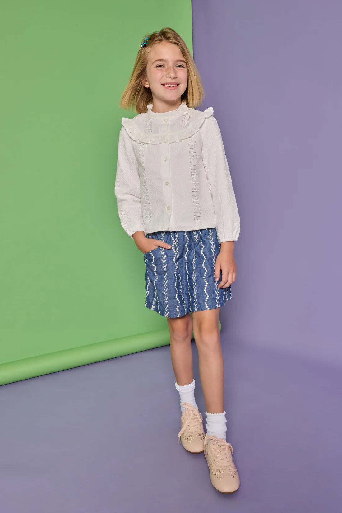 A young girl stands against a split green and purple studio backdrop, wearing a white long-sleeve blouse with a ruffled high collar and a blue patterned skirt.