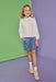 A young girl stands against a split green and purple studio backdrop, wearing a white long-sleeve blouse with a ruffled high collar and a blue patterned skirt.