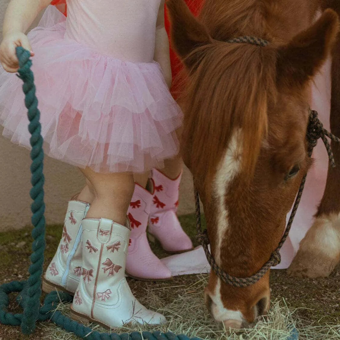 Child in pink dress and cowboy boots standing next to a horse