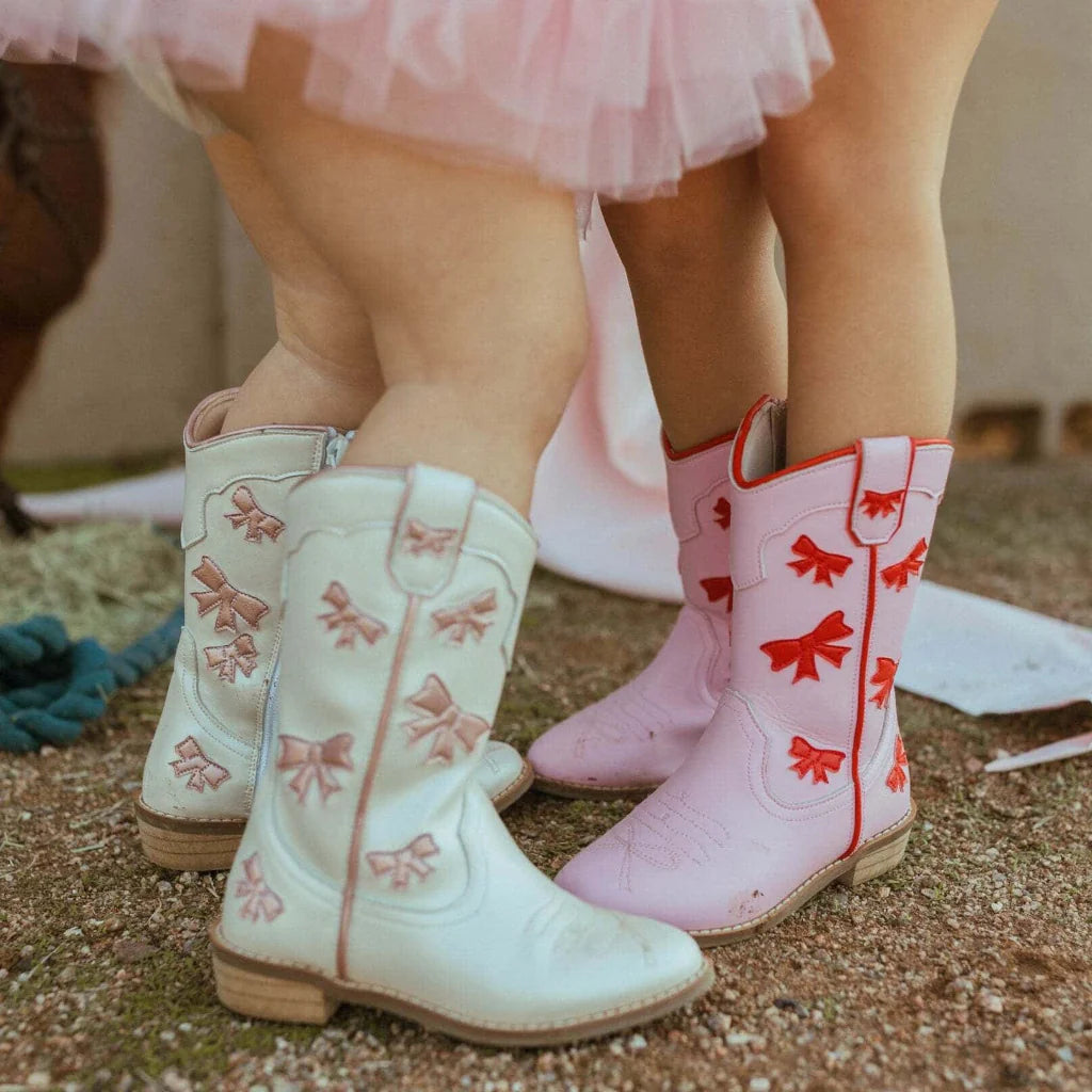 Children's white and pink cowboy boots with bow designs on a sandy ground.