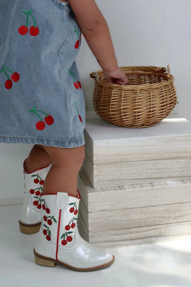Child wearing a denim dress with cherry pattern and white boots with red cherries, standing next to a woven basket.