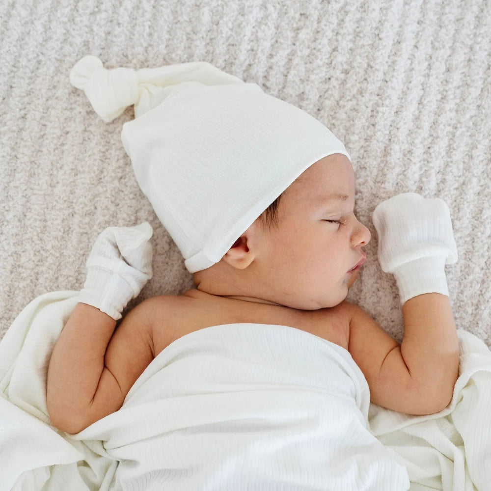 A sleeping newborn baby wearing a white knit beanie hat and matching white mittens, swaddled in a white blanket and lying on a textured cream blanket.