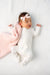 Newborn baby in a white onesie with a pink bow, lying on a white textured surface.
