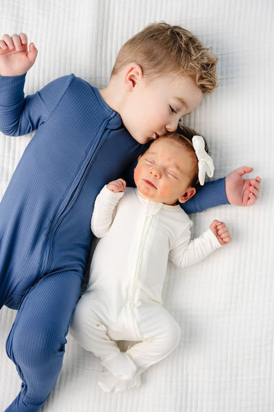 Child in a blue outfit lying next to a baby in a white outfit on a white blanket.