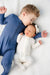 Child in a blue outfit lying next to a baby in a white outfit on a white blanket.