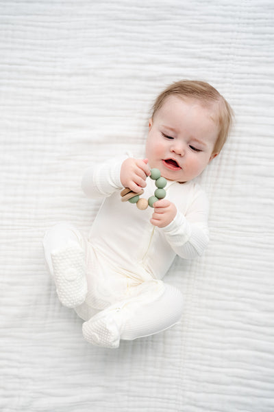 A baby wearing a white footed onesie lies on a white textured blanket, holding a green wooden bead teether.