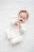 A baby wearing a white footed onesie lies on a white textured blanket, holding a green wooden bead teether.