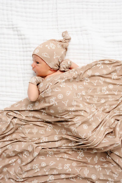Baby wrapped in a beige blanket with cowboy pattern, wearing a matching hat, on a white background.