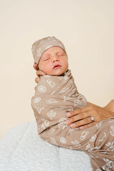 Newborn baby swaddled in a brown blanket with cowboy pattern, wearing a matching headband, held by a person against a beige background.