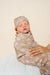 Newborn baby swaddled in a brown blanket with cowboy pattern, wearing a matching headband, held by a person against a beige background.