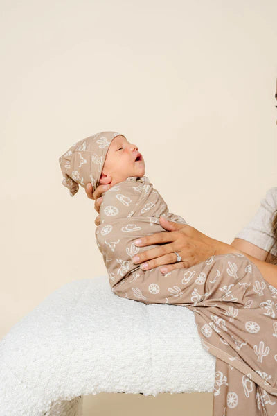 Baby wrapped in a patterned swaddle and cap, held by a person against a plain background