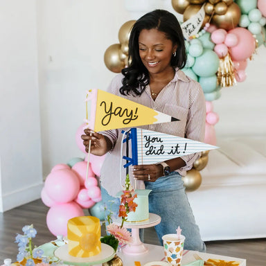 Woman holding 'Yay!' and 'You did it!' signs in a celebratory setting with balloons and cake.